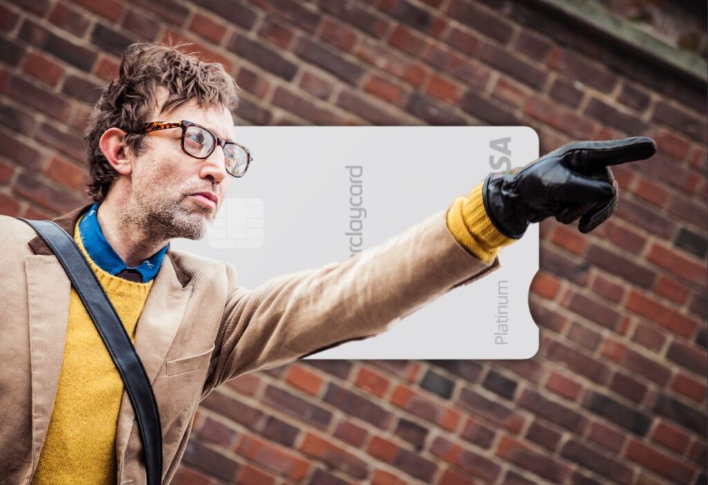 A man in a beige blazer and glasses gestures forward, standing in front of a brick wall with a Barclaycard Platinum Balance Transfer Card in the background.