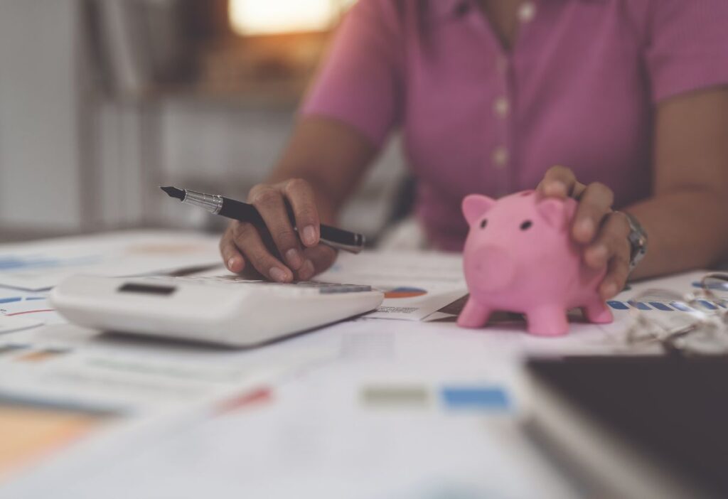 Person reviewing financial documents at a desk, holding a piggy bank and using a calculator, symbolising budgeting and saving.