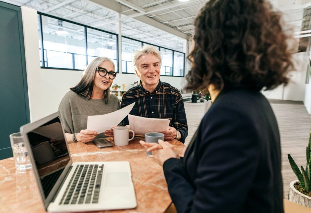 A mature couple discussing life insurance plans with a financial advisor, sitting at a table with documents and a laptop.