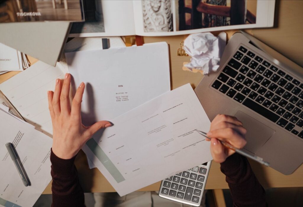 Close-up of hands reviewing paperwork on a cluttered desk, with a laptop, pen, and calculator, suggesting a focus on financial analysis.