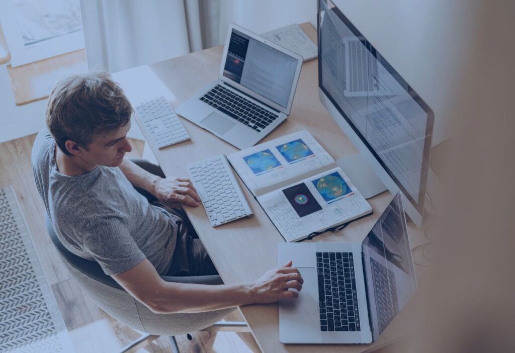 Man working on multiple devices at a desk, surrounded by notebooks and screens, representing a focused effort on managing financial data securely.